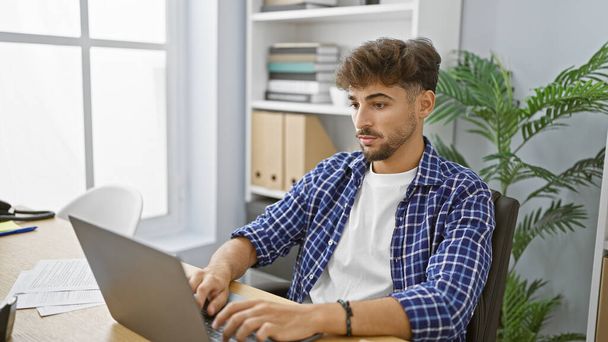Yakışıklı genç Arap adam, rahat ama ciddi ve zarif bir iş adamı, kapalı ofis alanında sıkı çalışıyor, başarı için dizüstü bilgisayarına odaklanıyor. - Fotoğraf, Görsel