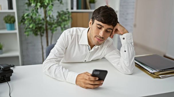 Joven hispano agotado, un atractivo profesional, aburrido y cansado en el trabajo, mirando la pantalla de su smartphone, sintiéndose atrapado dentro de la oficina - Foto, imagen