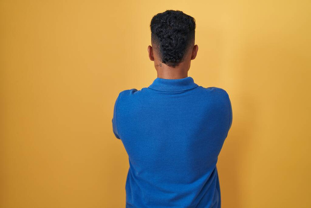 Jeune homme hispanique debout sur fond jaune debout vers l'arrière regardant loin avec les bras croisés  - Photo, image