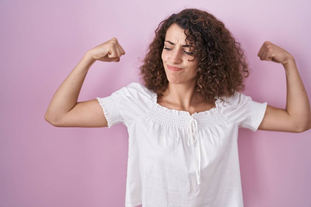 Hispanic woman with curly hair standing over pink background showing arms muscles smiling proud. fitness concept.  - Zdjęcie, obraz