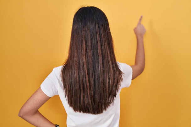 Young beautiful woman standing over yellow background posing backwards pointing ahead with finger hand  - Photo, Image
