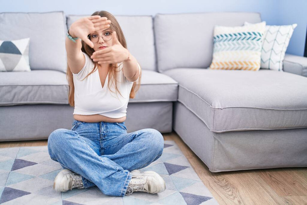 Mujer caucásica joven sentada en el suelo en la sala de estar haciendo marco usando las manos palmas y los dedos, perspectiva de la cámara  - Foto, imagen