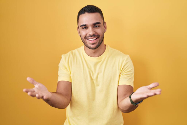Young hispanic man standing over yellow background smiling cheerful offering hands giving assistance and acceptance.  - Фото, зображення