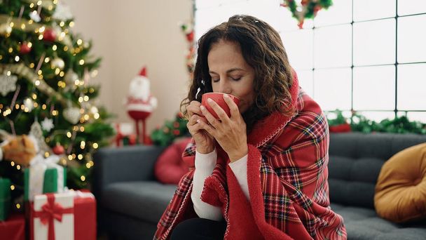 Mujer hispana de mediana edad bebiendo café sentada en el sofá junto al árbol de Navidad en casa - Foto, Imagen