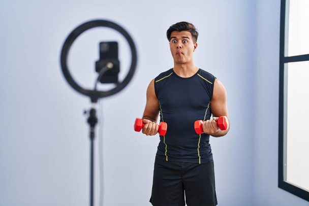 Hispanic man recording training tutorial at the gym making fish face with mouth and squinting eyes, crazy and comical.  - Photo, Image