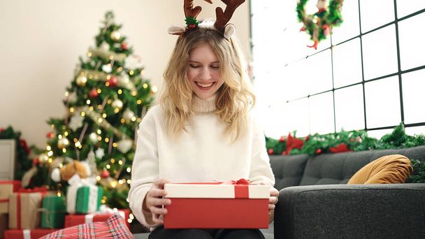 Joven mujer rubia desembalaje regalo sentado en el sofá por el árbol de Navidad en casa - Foto, imagen