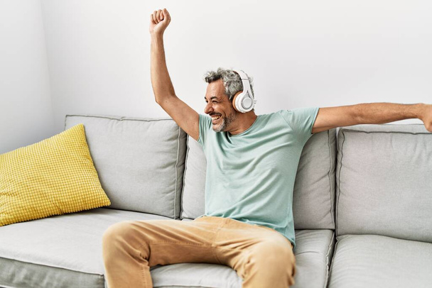 Седовласый мужчина средних лет танцует и слушает музыку, сидя дома на диване - Фото, изображение