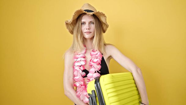 Giovane donna bionda turista indossa bikini e cappello estivo tenendo valigia su isolato sfondo giallo - Foto, immagini