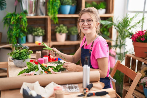 Young blonde woman florist make bouquet of flowers sitting on table at flower shop - Photo, Image
