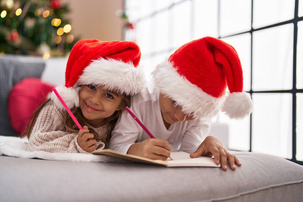 Брат и сестра пишут Санта-Клаусу письмо лежа на диване у рождественской елки дома - Фото, изображение