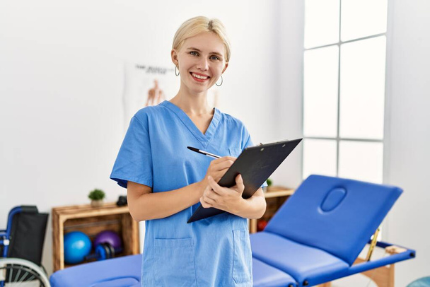 Young blonde woman pysiotherapist smiling confident writing on document at rehab clinic - 写真・画像