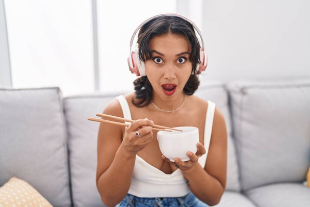 Nuori latino nainen syö aasialaista ruokaa käyttäen syömäpuikkoja shokki kasvot, etsivät skeptinen ja sarkastinen, yllättynyt suu auki  - Valokuva, kuva