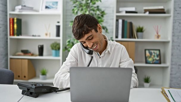 Itsevarma latinoalan liikemies, nuoren miehen hymyilevä muotokuva, kun hän puhuu puhelimessa, työstää läppäriään toimiston pöydällä, hukuttaen menestystä - Valokuva, kuva