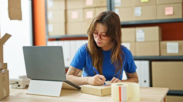 Junge schöne hispanische Frau E-Commerce-Unternehmer mit Laptop schreiben auf Paket im Büro - Foto, Bild