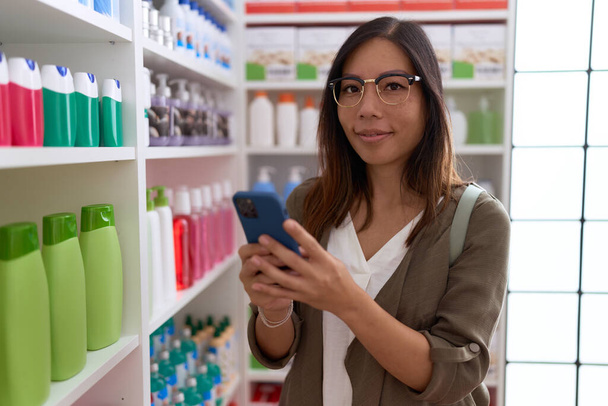 Joven asiática mujer cliente sonriendo confiado usando smartphone en farmacia - Foto, Imagen