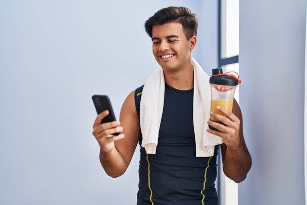 Jovem hispânico usando o smoothie de proteína de consumo de smartphones no centro esportivo - Foto, Imagem