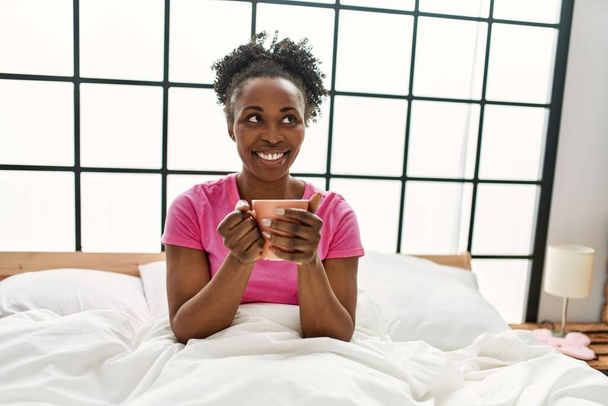 Afrikaans amerikaanse vrouw drinken kopje koffie zitten op bed in de slaapkamer - Foto, afbeelding