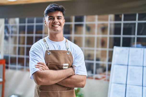 Молодий іспаномовний чоловік офіціант, усміхнений впевнено стоїть зі зброєю перетнув жест на терасі кав'ярні
 - Фото, зображення