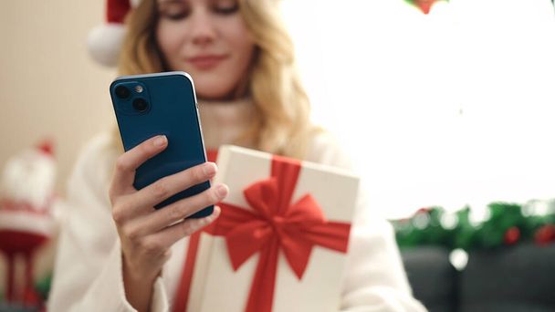 jong blond vrouw met behulp van smartphone holding christmas gift thuis - Foto, afbeelding