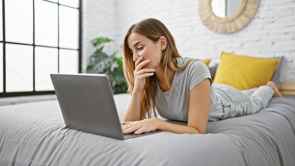 Houkutteleva nuori blondi nainen makaa mukavasti sängyssä ja käyttää kannettavaa tietokonettaan hypätä verkossa viihtyisässä kotiympäristössä, jossa on rento ilme, joka tuo elämän makuuhuoneeseensa. - Valokuva, kuva