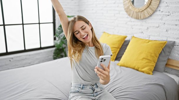 Joven mujer caucásica alegre, una rubia atractiva, celebrando una victoria mientras envía mensajes de texto en su teléfono inteligente, cómodamente sentada en una cama en su acogedor dormitorio - Foto, imagen