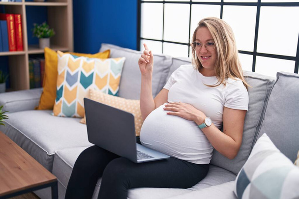 Νεαρή έγκυος γυναίκα κάθεται στον καναπέ στο σπίτι χρησιμοποιώντας φορητό υπολογιστή χαμογελώντας χαρούμενος δείχνοντας με το χέρι και το δάχτυλο στο πλάι  - Φωτογραφία, εικόνα