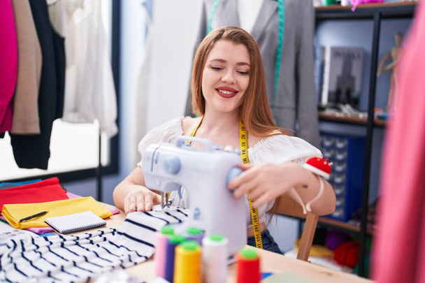 Молодая рыжая женщина портной улыбается уверенно с помощью швейной машинки на швейной фабрике - Фото, изображение