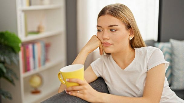 Genç sarışın kadın evdeki kanepede kahve içiyor. - Fotoğraf, Görsel
