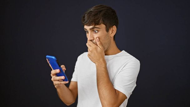 Молодой латиноамериканец смотрит на смартфон с удивленным выражением лица на изолированном черном фоне - Фото, изображение