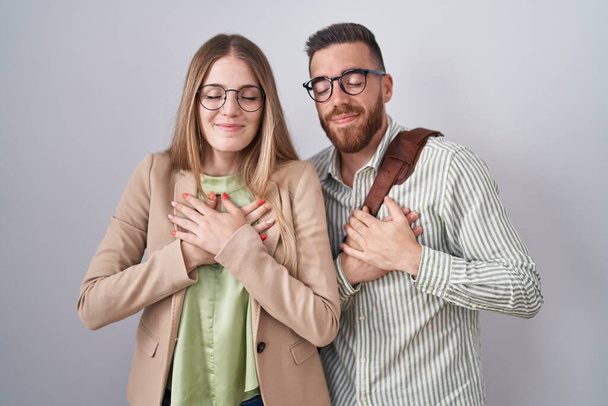 Ein junges Paar steht vor weißem Hintergrund und lächelt mit geschlossenen Augen und dankbarer Geste auf der Brust. Gesundheitskonzept.  - Foto, Bild