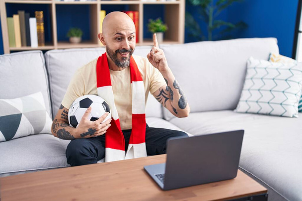 Spanyol férfi tetoválásokkal nézi a focimeccset huligán kezében labdát a laptopon meglepve egy ötlettel vagy kérdéssel ujjal mutogatva boldog arccal, egyes számú  - Fotó, kép