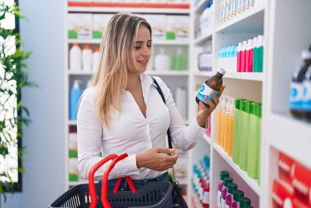 Young blonde woman customer holding medication bottle of shelving at pharmacy - Photo, Image