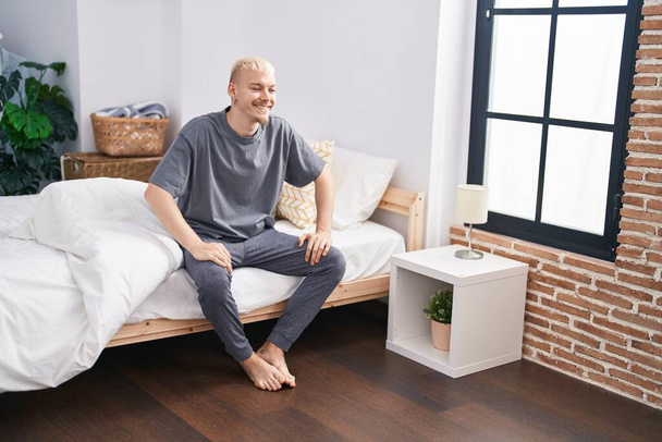 Nuori valkoihoinen mies hymyilee luottavainen istuu sängyllä makuuhuoneessa - Valokuva, kuva