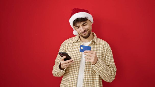 孤立した赤い背景上のスマートフォンでクリスマスオンラインショッピングをしている若いアラブの男 - 写真・画像