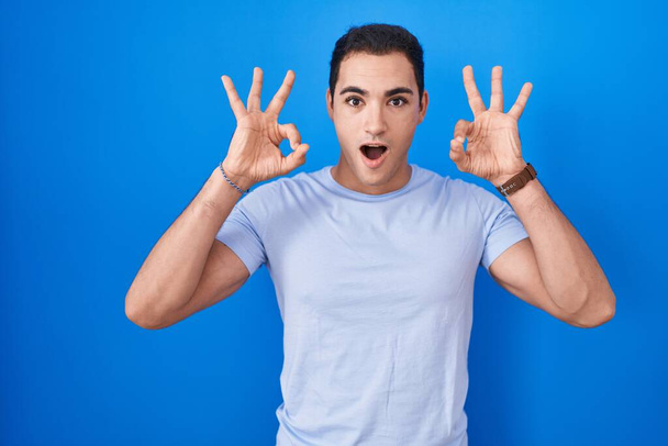 Νεαρός Ισπανόφωνος στέκεται πάνω από το μπλε φόντο φαίνεται έκπληκτος και σοκαρισμένος κάνει εντάξει σύμβολο έγκρισης με τα δάχτυλα. τρελή έκφραση  - Φωτογραφία, εικόνα