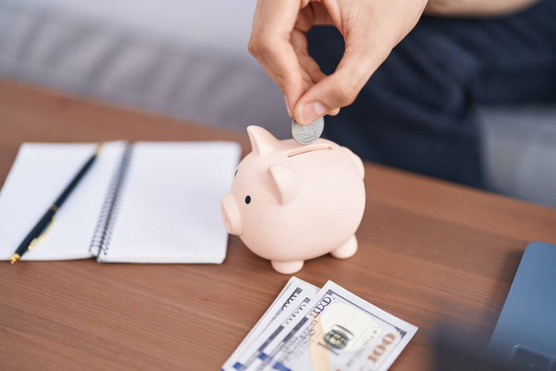 Young blond man inserting coin on piggy bank at home - Photo, Image