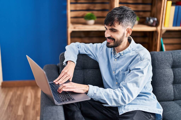 Молодий іспаномовний чоловік використовує ноутбук, сидячи на дивані вдома
 - Фото, зображення