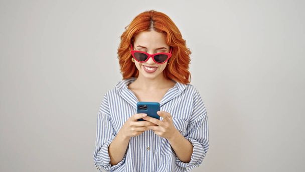 Genç kızıl saçlı, güneş gözlüğü takan, izole edilmiş beyaz arka plan üzerinde akıllı telefon kullanan bir kadın. - Fotoğraf, Görsel