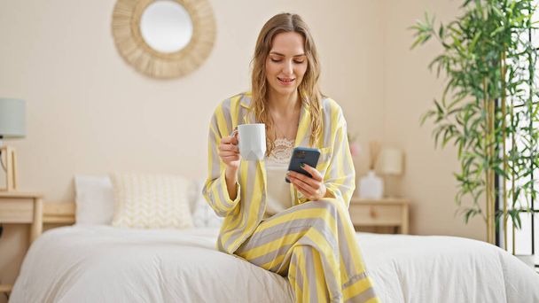Young blonde woman using smartphone drinking coffee at bedroom - Fotografie, Obrázek