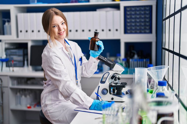 Νεαρή καυκάσια γυναίκα επιστήμονας γράψει στο έγγραφο κρατώντας μπουκάλι στο εργαστήριο - Φωτογραφία, εικόνα