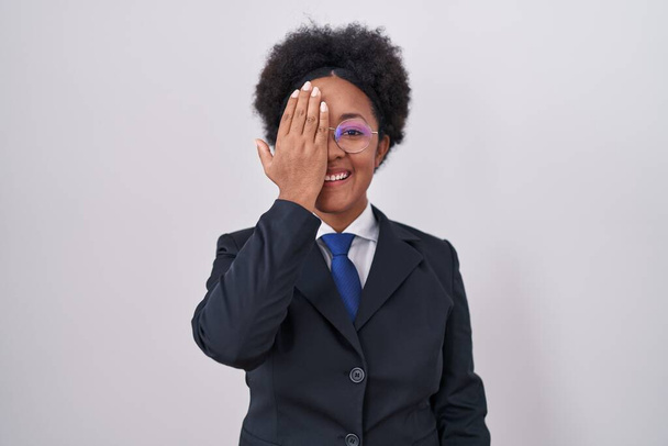 Beautiful african woman with curly hair wearing business jacket and glasses covering one eye with hand, confident smile on face and surprise emotion.  - Fotografie, Obrázek