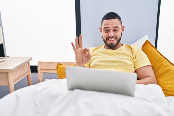 Ισπανόφωνος άνθρωπος χρησιμοποιώντας φορητό υπολογιστή στο κρεβάτι κάνει ok υπογράψει με τα δάχτυλα, χαμογελώντας φιλικό gesturing εξαιρετικό σύμβολο  - Φωτογραφία, εικόνα