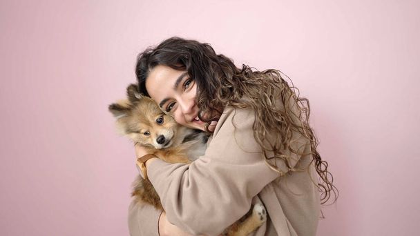 Young hispanic woman with dog smiling confident standing over isolated pink background - 写真・画像