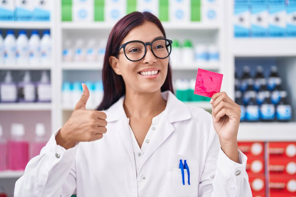 Junge hispanische Frau, die in der Apotheke arbeitet, hält Kondom und lächelt glücklich und positiv, Daumen hoch und Zeichen der Anerkennung  - Foto, Bild