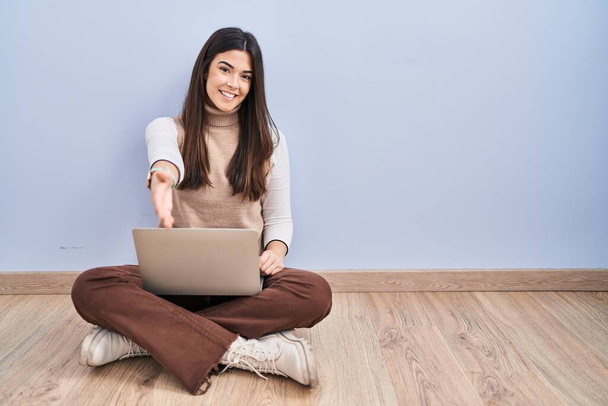 Mladá bruneta žena pracující pomocí notebooku sedí na podlaze s úsměvem přátelské nabízí potřesení rukou jako pozdrav a přivítání. úspěšný obchod.  - Fotografie, Obrázek