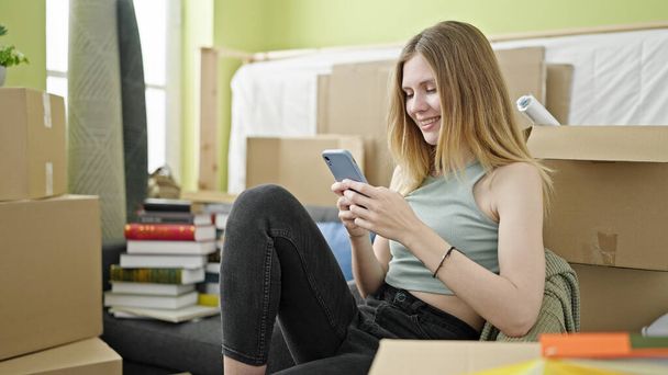 Young blonde woman using smartphone sitting on sofa at new home - Fotoğraf, Görsel