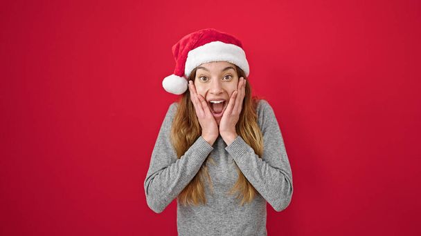 若い白人女性は隔離された赤い背景の上にクリスマスの帽子を身に着けている驚きの表情 - 写真・画像