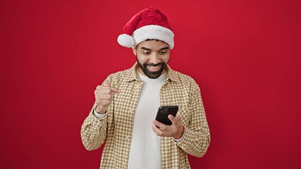 Young hispanic man wearing christmas hat pointing to smartphone over isolated red background - Photo, Image