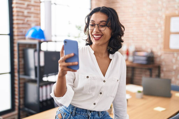 Young hispanic woman working at the office using smartphone looking positive and happy standing and smiling with a confident smile showing teeth  - Fotoğraf, Görsel