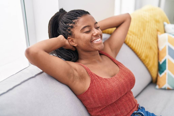 Mujer afroamericana relajada con las manos en la cabeza sentada en el sofá en casa - Foto, imagen
